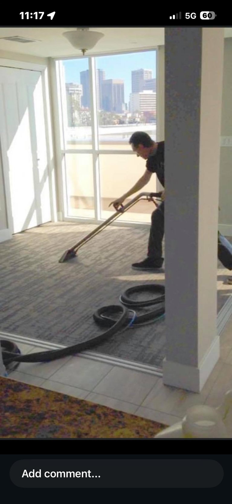 Man steam cleaning a carpeted floor in a high-rise apartment with city skyline view through glass doors